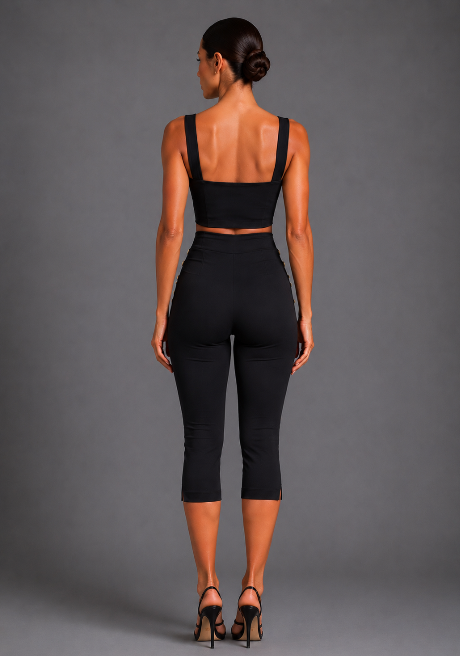 Back image of a model wearing the VSPR Athena Square Neckline Button Detail Black Crop Top with Capri Cropped Black Button Detail Trousers, displaying the sleek fitted back, wide straps, and cropped tailored design against a grey studio backdrop.
