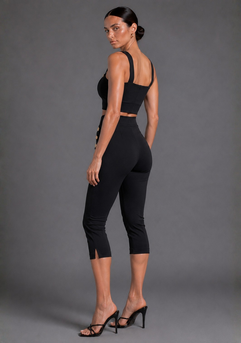 A woman modeling a black two-piece activewear set consisting of a strappy crop top and high-waisted capri leggings with side cutouts, paired with black strappy high-heeled sandals, against a solid gray studio background.