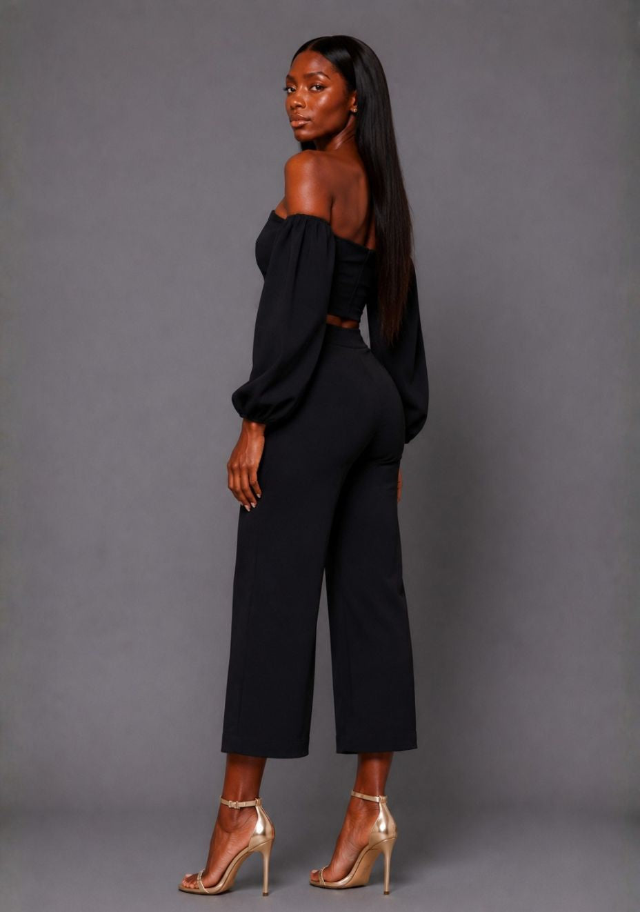 Side back image of a model wearing the VSPR Mindi Wide Leg Black Cropped Trousers, showcasing the smooth high-rise fit and flowing wide-leg shape against a grey studio backdrop.