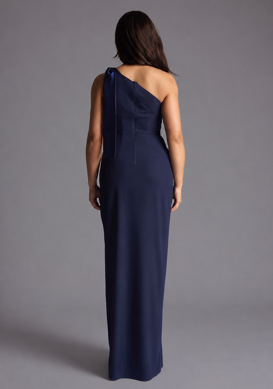 Woman wearing the Vesper Petite Olympia navy one shoulder satin contrast maxi dress against a grey background, back view