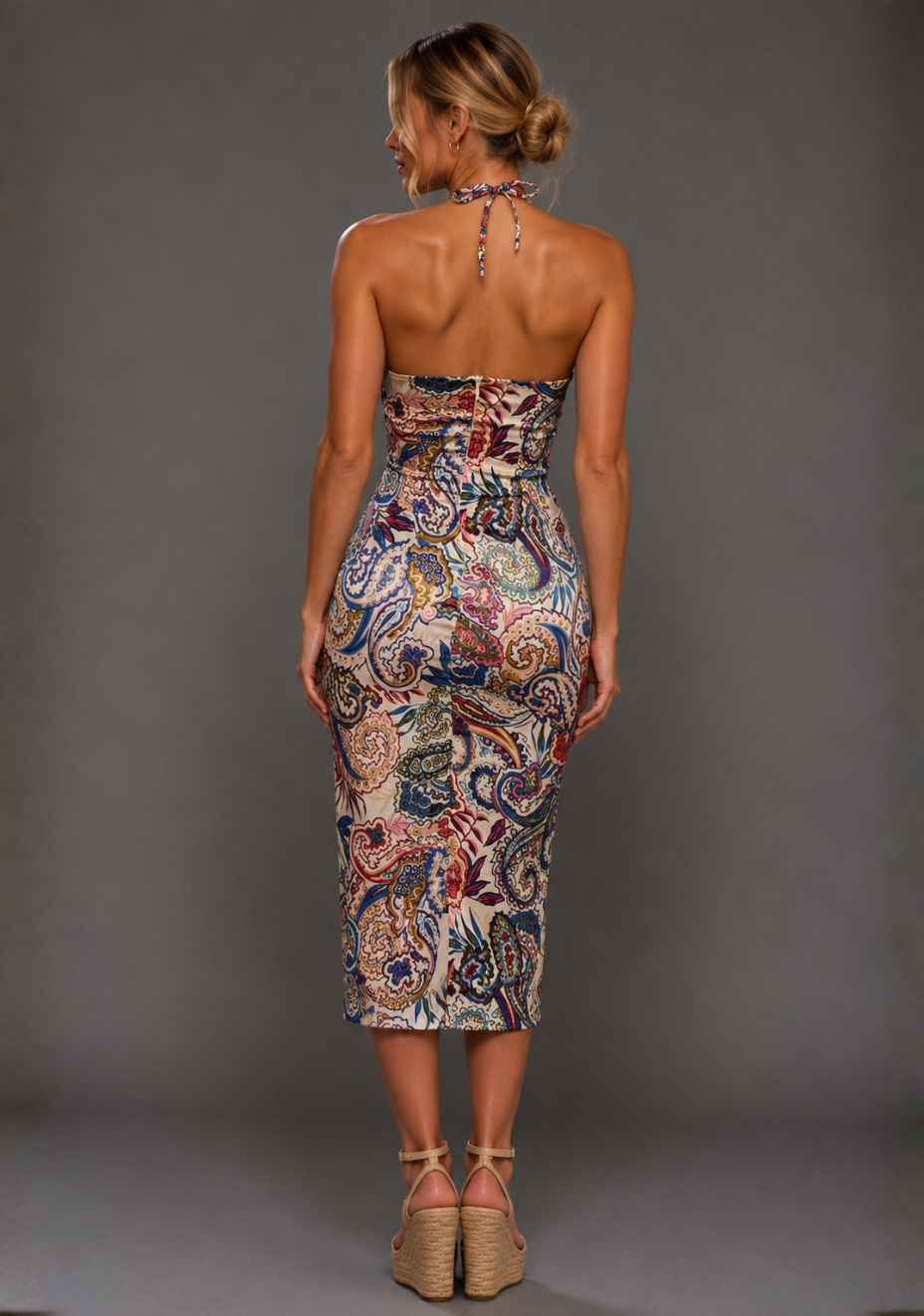 A woman viewed from the back wearing a strapless halter midi dress with a dense paisley pattern in red, blue, beige, and white, paired with beige wedge sandals against a solid gray studio background.