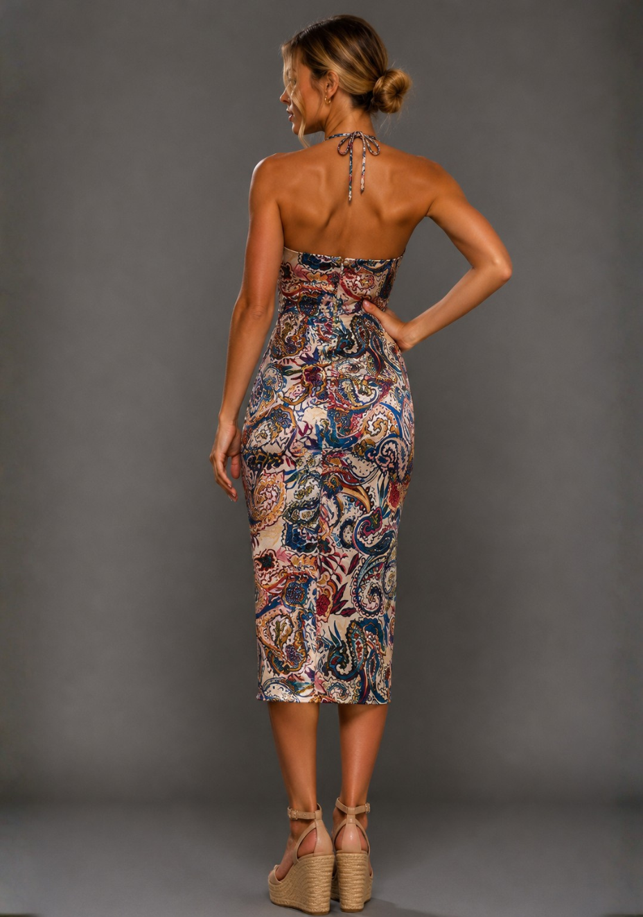 A woman modeling a sleeveless halter midi dress with a colorful paisley print, shown from the back against a solid gray studio background.