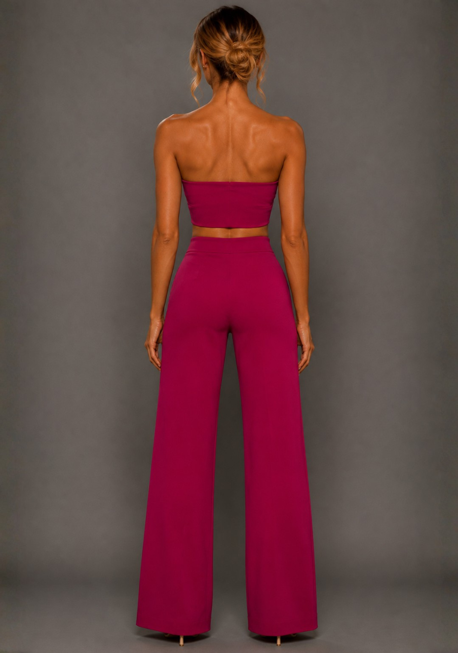 Back image of a model wearing the VSPR Renata Strapless Magenta Crop Top and matching VSPR Marty Magenta Wide Leg Trousers, revealing the straight strapless back and wide-leg silhouette, standing against a grey studio backdrop.