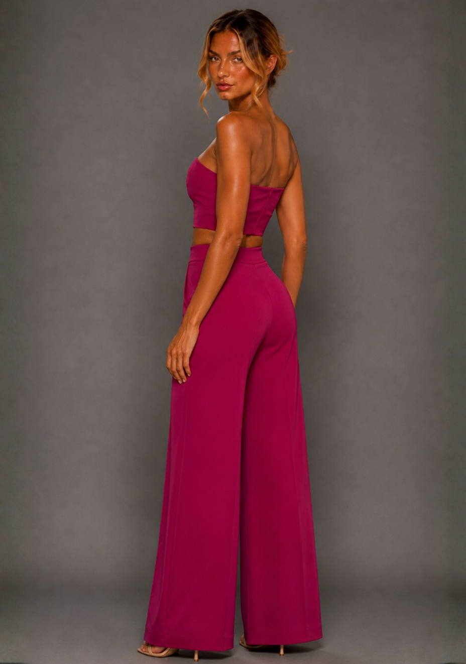 Side back image of a model wearing the VSPR Marty Magenta Wide Leg Pants, showcasing the high-rise waist and sweeping wide leg, paired with the VSPR Renata Strapless Magenta Crop Top, standing against a grey studio backdrop.