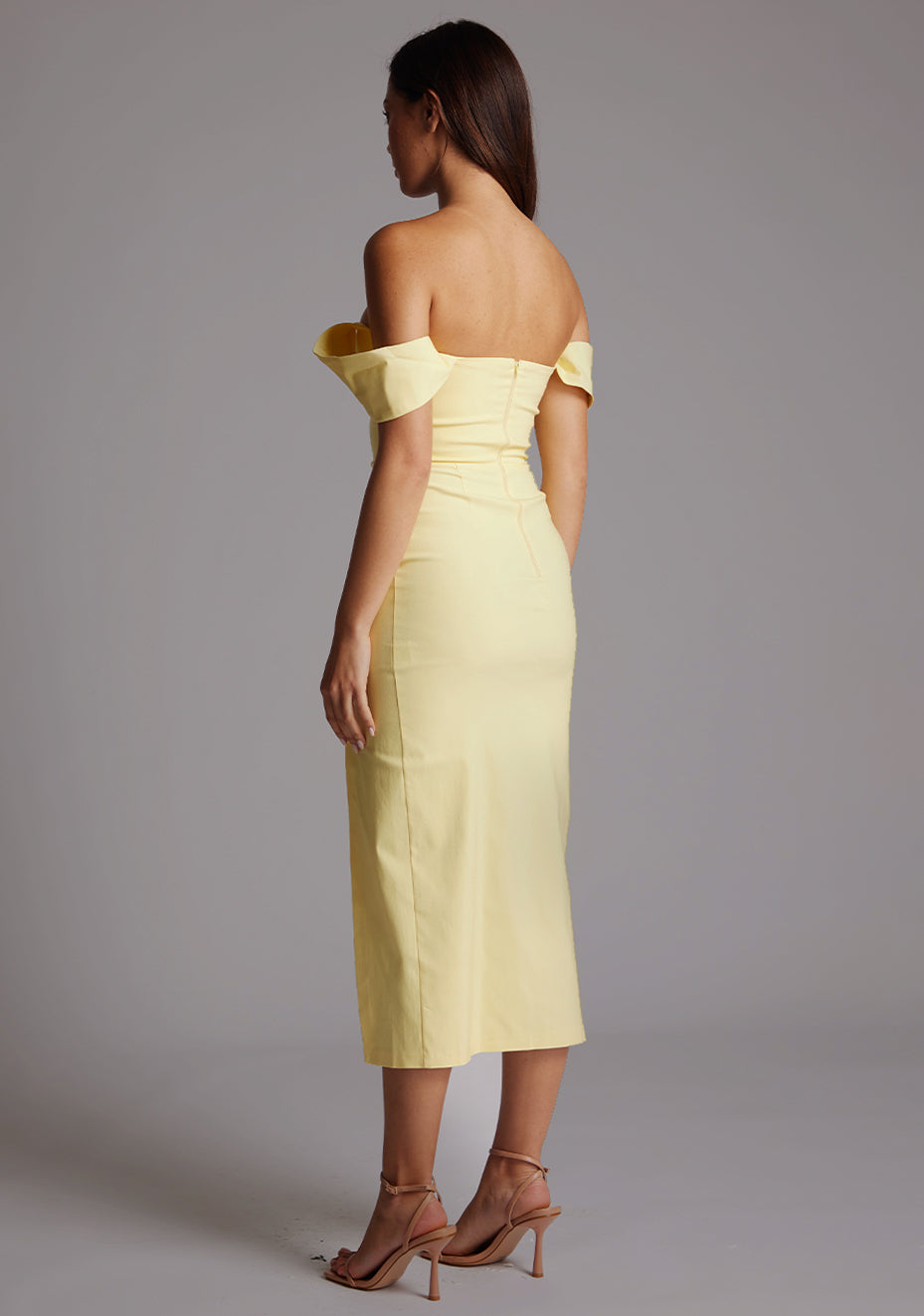 Side back image of a model wearing the VSPR Astra Bardot Lemon Midaxi Dress, highlighting its fitted shape, midaxi length, and smooth super-stretch fabric against a grey studio backdrop.