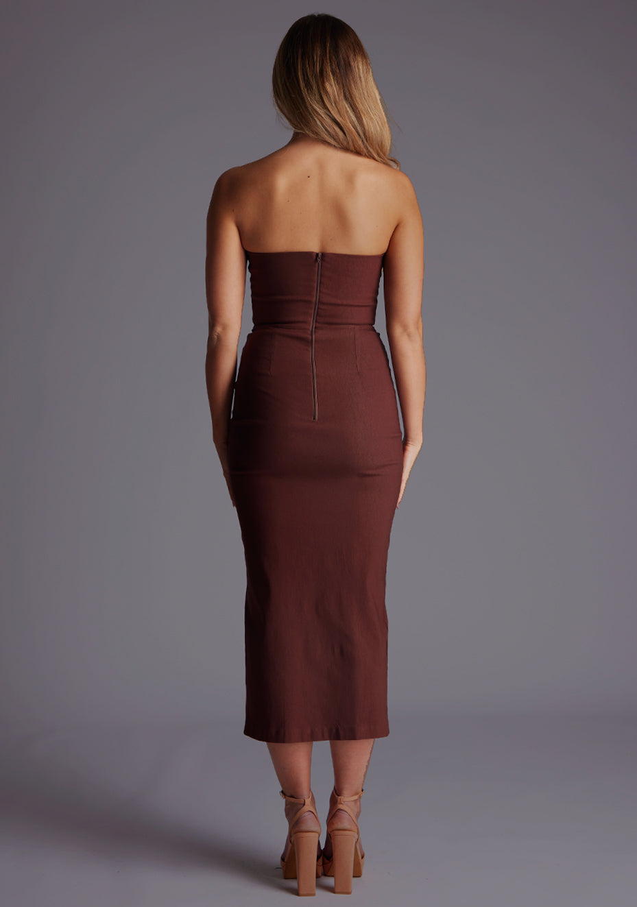 Back image of a blonde model wearing a chocolate midaxi dress with a strapless gown, front cut-out that adds a touch of allure, while the front skirt split adds a modern flair. The design featured is the Vesper Delaney Strapless V-Front Midaxi Dress Chocolate..