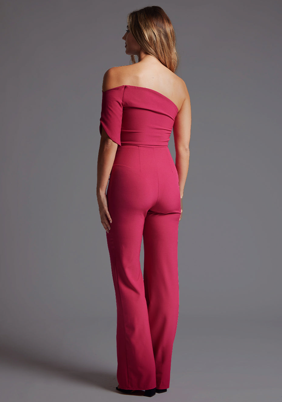 Side back image of a model wearing a with a stunning one-shoulder design and a body-hugging fit that accentuates your curves in all the right places and side zip fastening ensures a seamless look. The design featured is the Vesper Ines One Shoulder Jumpsuit in Raspberry.