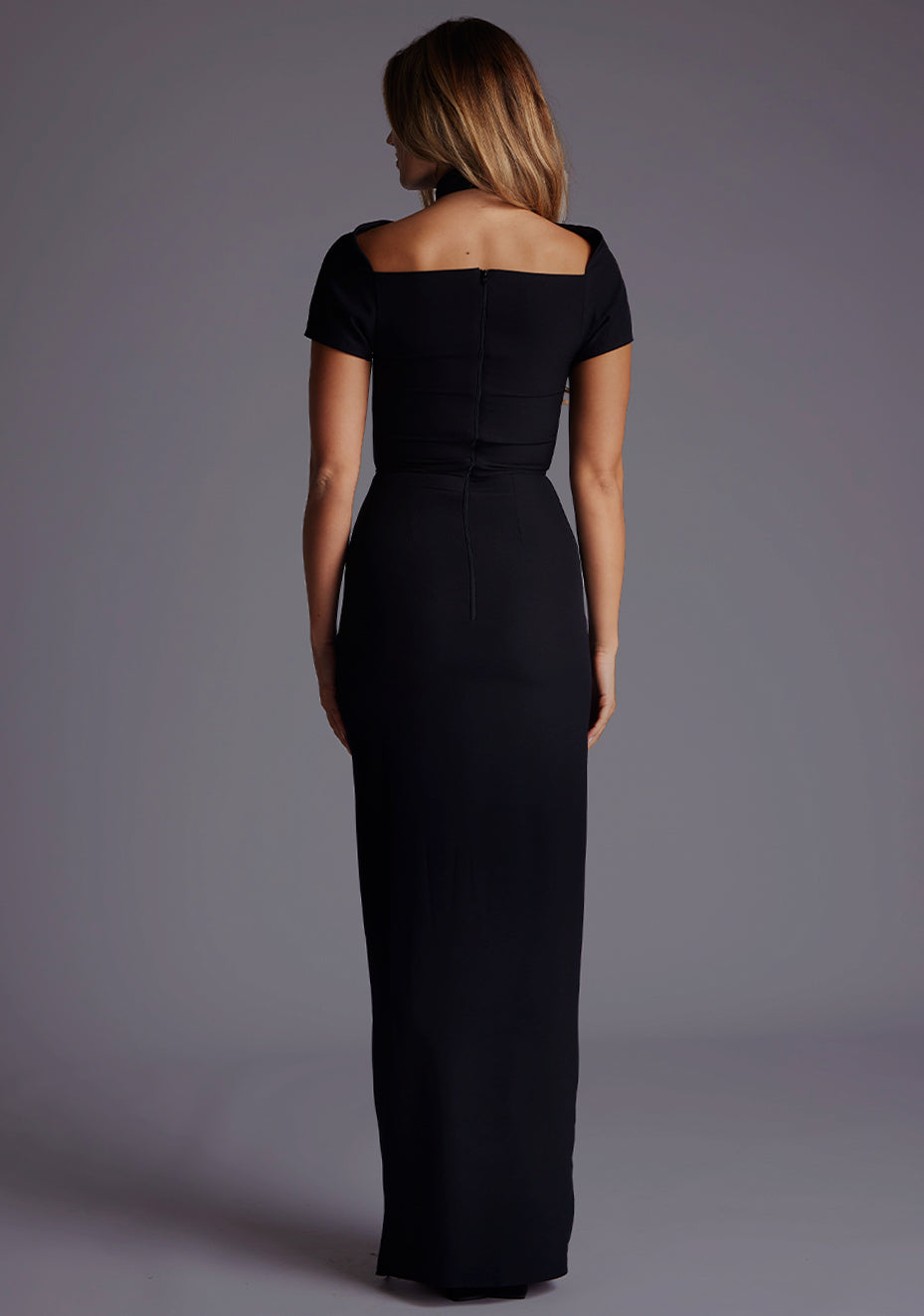 Back image of a blonde model wearing a Black Maxi Dress with a high neck, with a subtle cut-out detail, and a front skirt split. The dress is a design feature is the Vesper Josephine High Neck Short Sleeve Maxi Dress in Black.