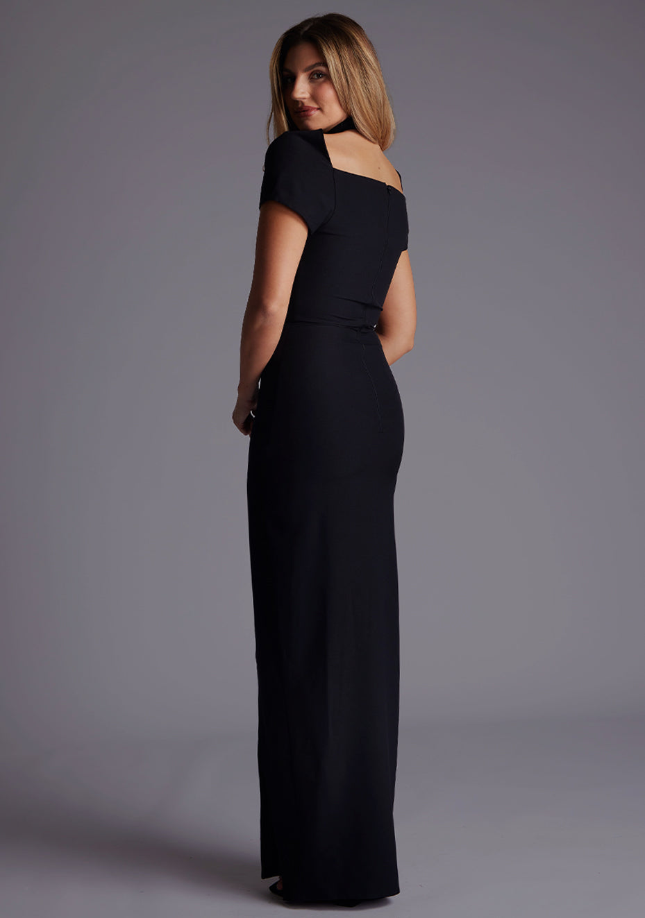 Side back image of a blonde model wearing a Black Maxi Dress with a high neck, with a subtle cut-out detail, and a front skirt split. The dress is a design feature is the Vesper Josephine High Neck Short Sleeve Maxi Dress in Black.