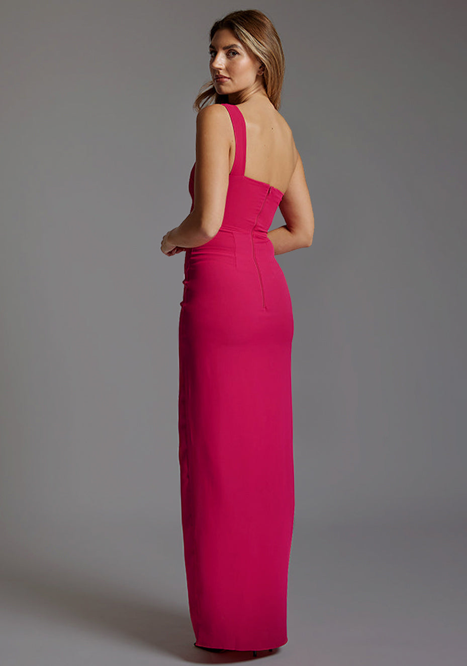 Back image of a blonde model wearing a ccerise maxi dress with a asymmetric neckline, front skirt split and beautiful cerise hue. The design featured is the Vesper Olivette Cerise Maxi Dress