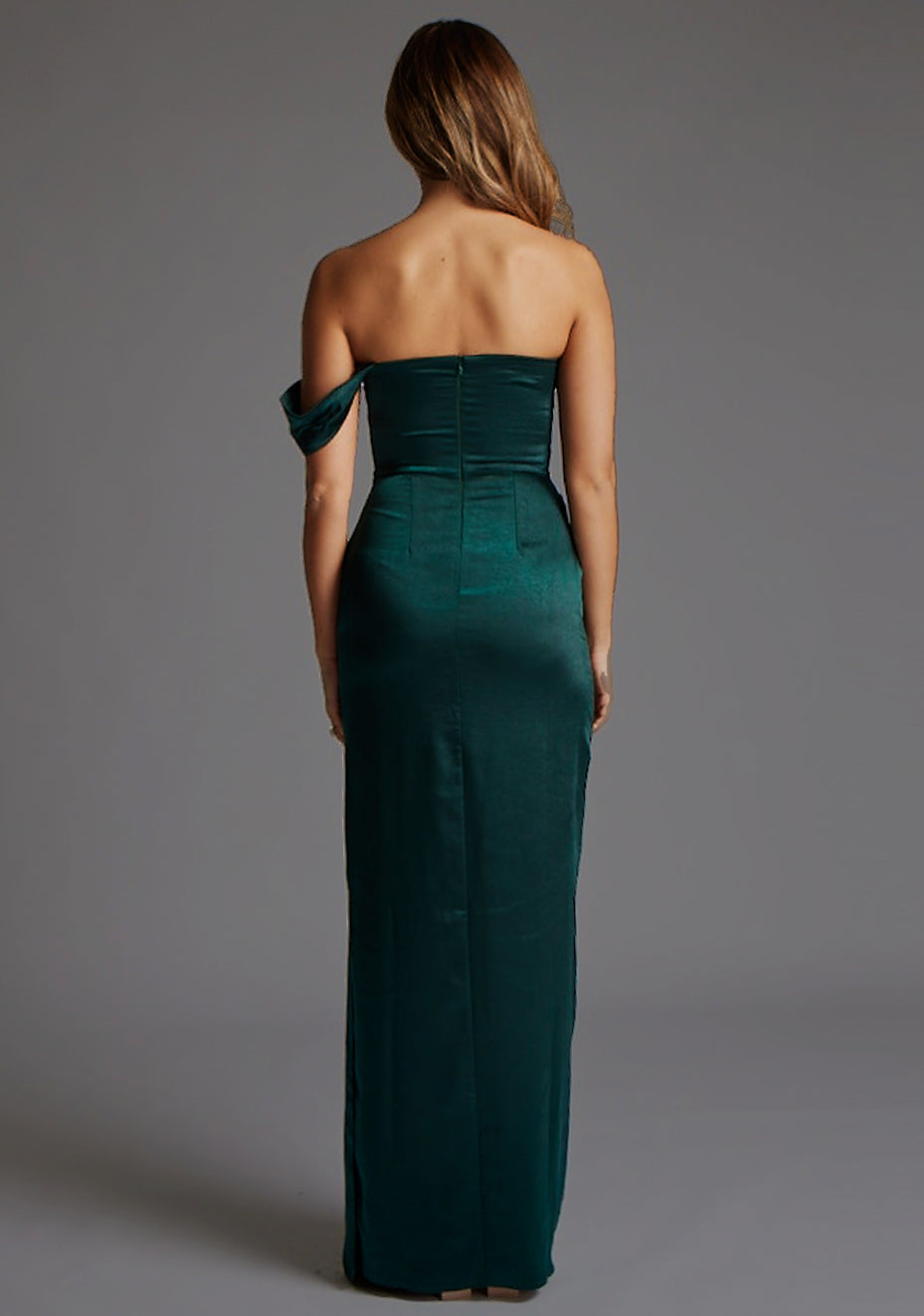 Back image of blonde model wearing a pine green maxi dress with a one-sleeve off-shoulder design, with a satin fabric. The design featured is the Vesper Roselie Pine Maxi Dress