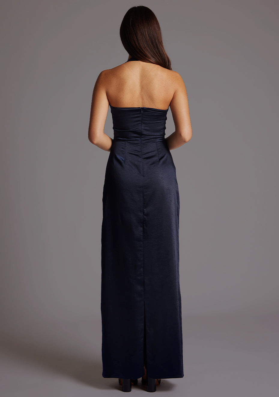 Back image of a model wearing a navy satin maxi dress with a halter cowl neck and low backless, the luxurious satin fabric slim-fit dress effortlessly flatters your figure. The design featured is the Vesper Saige Navy Satin Maxi Dress.