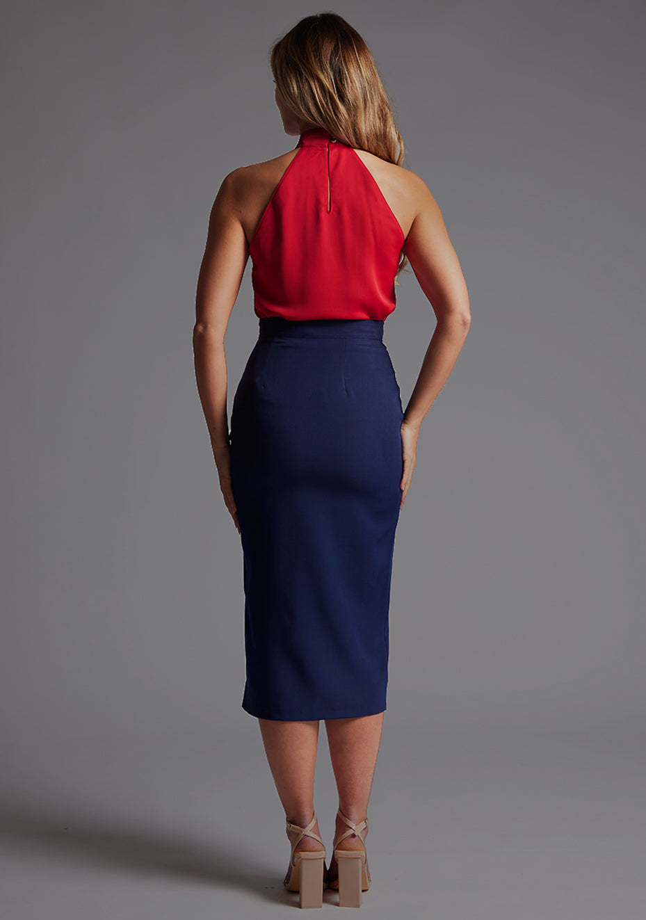 Back image of a model wearing a red top with a high neck, sleeveless with wide pleat detail at front with a centre back cut-out with statement gold buttons fastening at the back of the neck. The design featured is the Vesper Toren Red Top pair with Vesper Tana Navy Fitted Skirt
