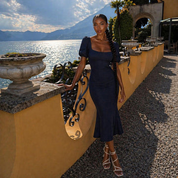 Image of a model wearing the vspr valencia fishtail midaxi dress in navy against a yellow wall overlooking the ocean