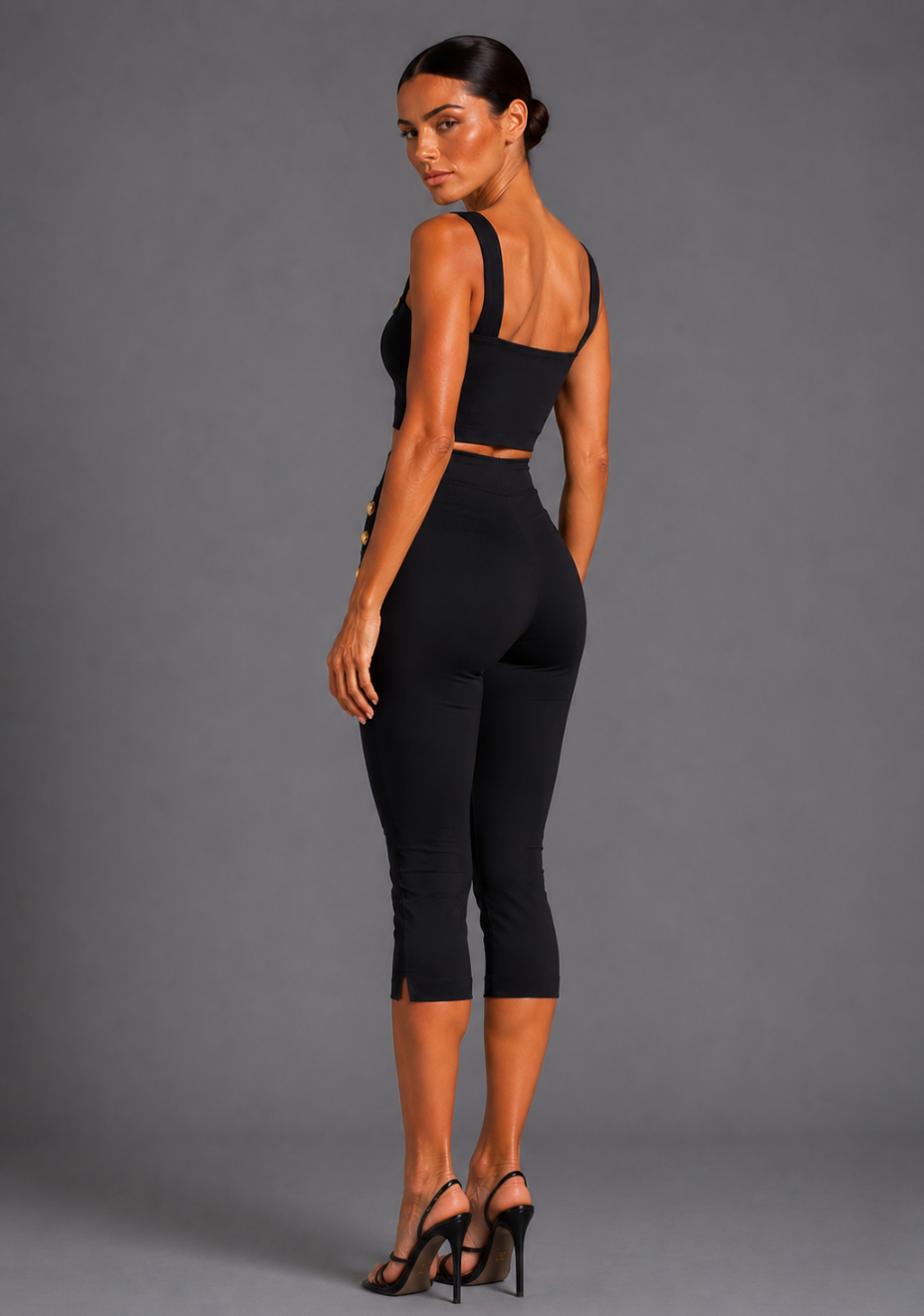 Side back image of a model wearing the VSPR Athena Square Neckline Button Detail Black Crop Top with Capri Cropped Black Button Detail Trousers, showcasing the clean tailored silhouette, structured fit, and cropped design, standing against a grey studio backdrop.
