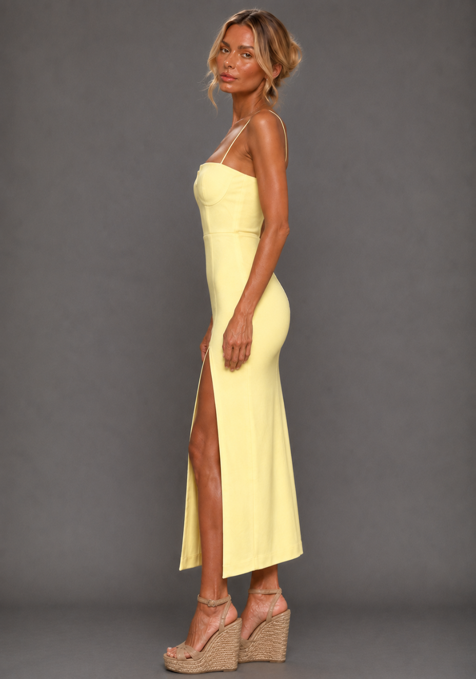 Side image of a model wearing the VSPR Aurora Spaghetti Strap Lemon Midaxi Dress in lemon, showing the body-skimming silhouette and front thigh split, standing against a grey studio backdrop.