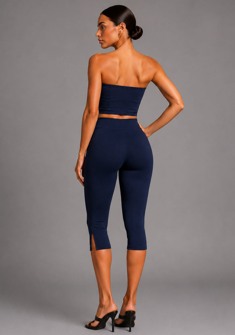 Side back image of a model wearing the VSPR Benedetta Strapless Navy Crop Top with Capri Navy Trousers, highlighting the clean strapless back, contoured silhouette, and tailored navy trousers against a grey studio backdrop.