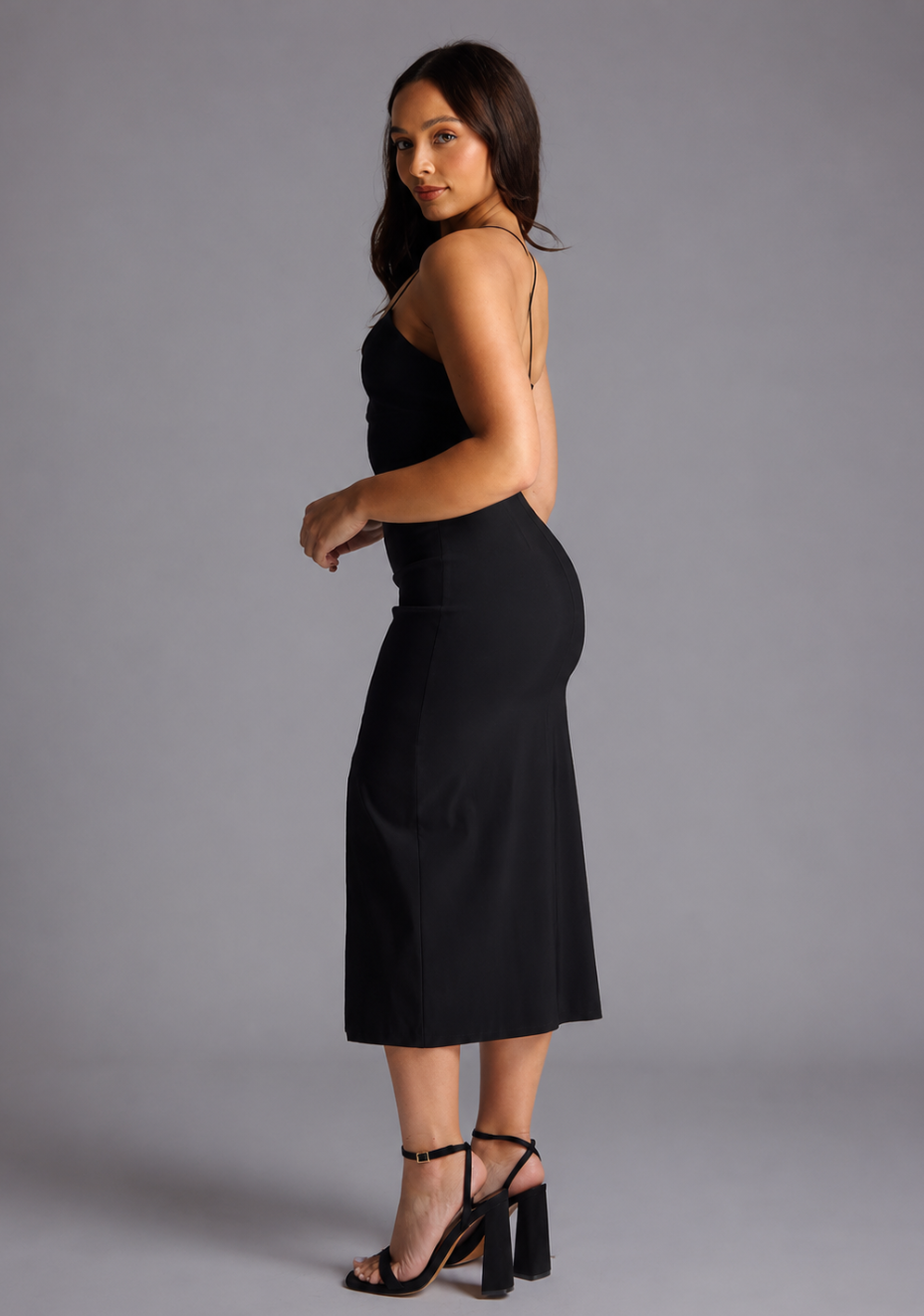 Side back image of a model wearing the VSPR Beryl Petite Strappy Bodycon Black Midaxi Dress in Black, showcasing its sleek figure-contouring shape and delicate crossover straps against a grey studio backdrop.