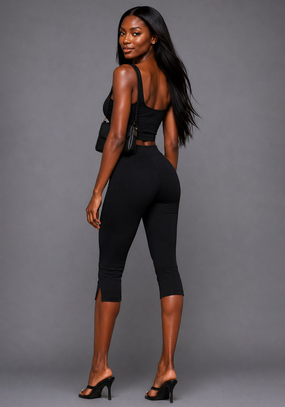 Side back image of a model wearing the VSPR Capri Cropped Black Trousers in Black, highlighting the sleek fit through the hips and cropped silhouette against a grey studio backdrop.