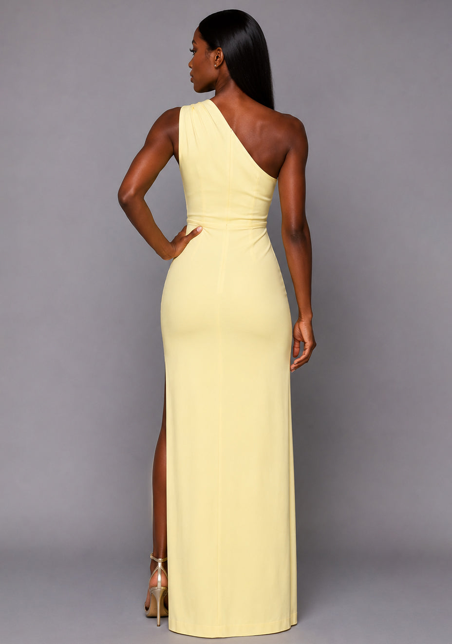 Back image of a model wearing the VSPR Charlotte One Shoulder Pleated Lemon Maxi Dress showcasing the clean asymmetric design and figure-skimming maxi silhouette against a grey studio backdrop.