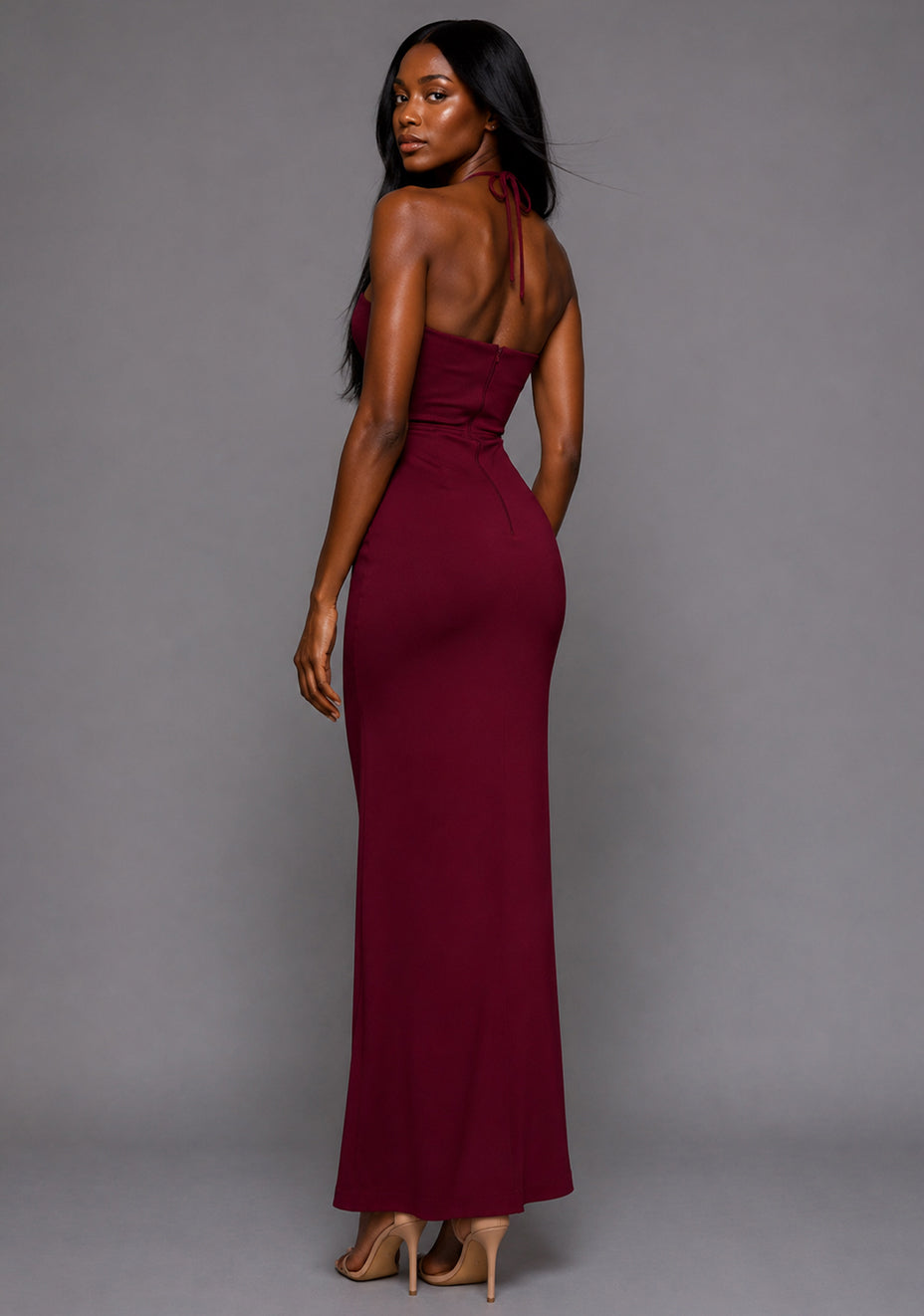 Side back image of a model wearing the VSPR Opal Cut-Out Halter Burgundy Maxi Dress, featuring the halter neckline, body-contouring silhouette, and fluid maxi skirt, photographed against a grey studio backdrop.