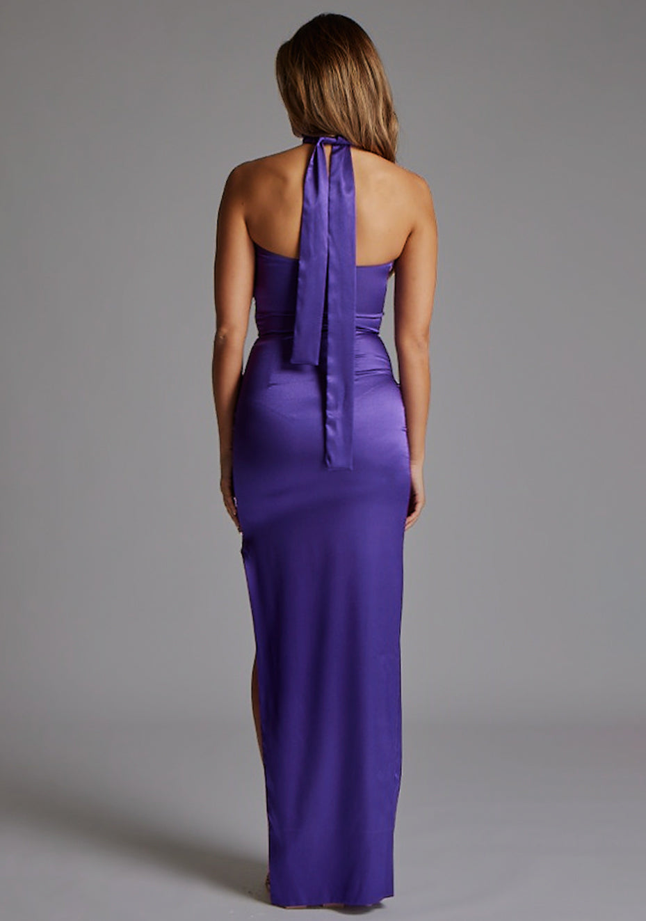 Back image of a model wearing the VSPR Bessie Satin Scarf Detail Purple Maxi Dress in Purple, showcasing the elegant halter straps, defined waist, and flowing maxi silhouette against a grey studio backdrop.