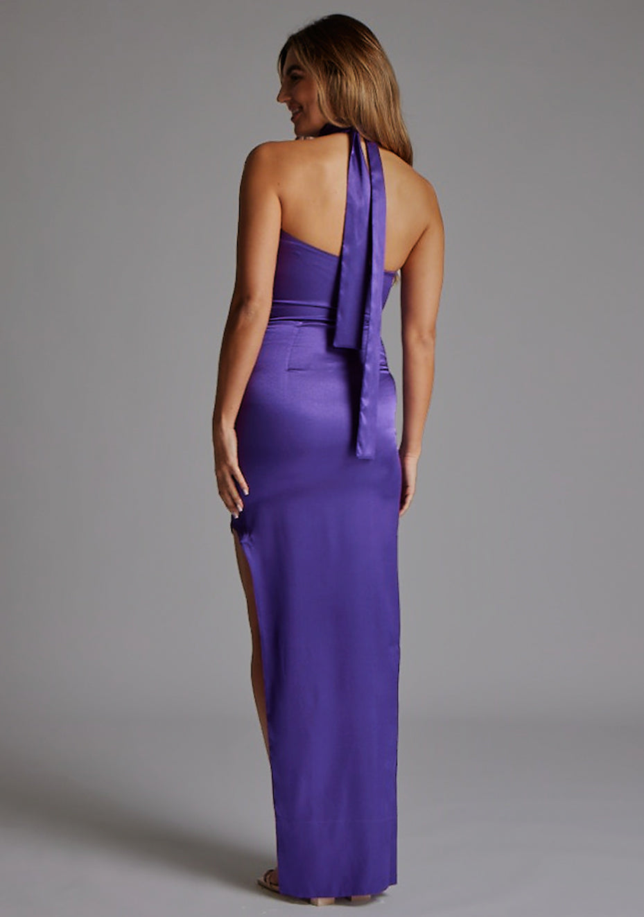 Side back image of a model wearing the VSPR Bessie Satin Scarf Detail Purple Maxi Dress, highlighting the lustrous satin fabric, halter straps, and smooth flowing silhouette against a grey studio backdrop.