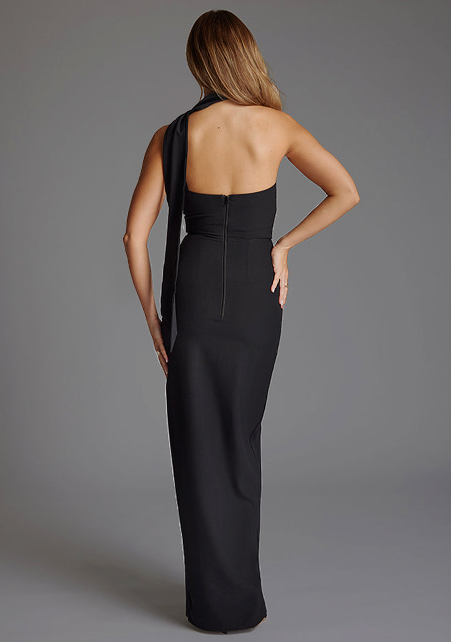 Back image of a model wearing a black maxi evening dress with asymmetric neckline, front skirt split and statement collar drape detail. The design featured is the Vesper Cordula Scarf Detail One Shoulder Maxi Dress  Black.