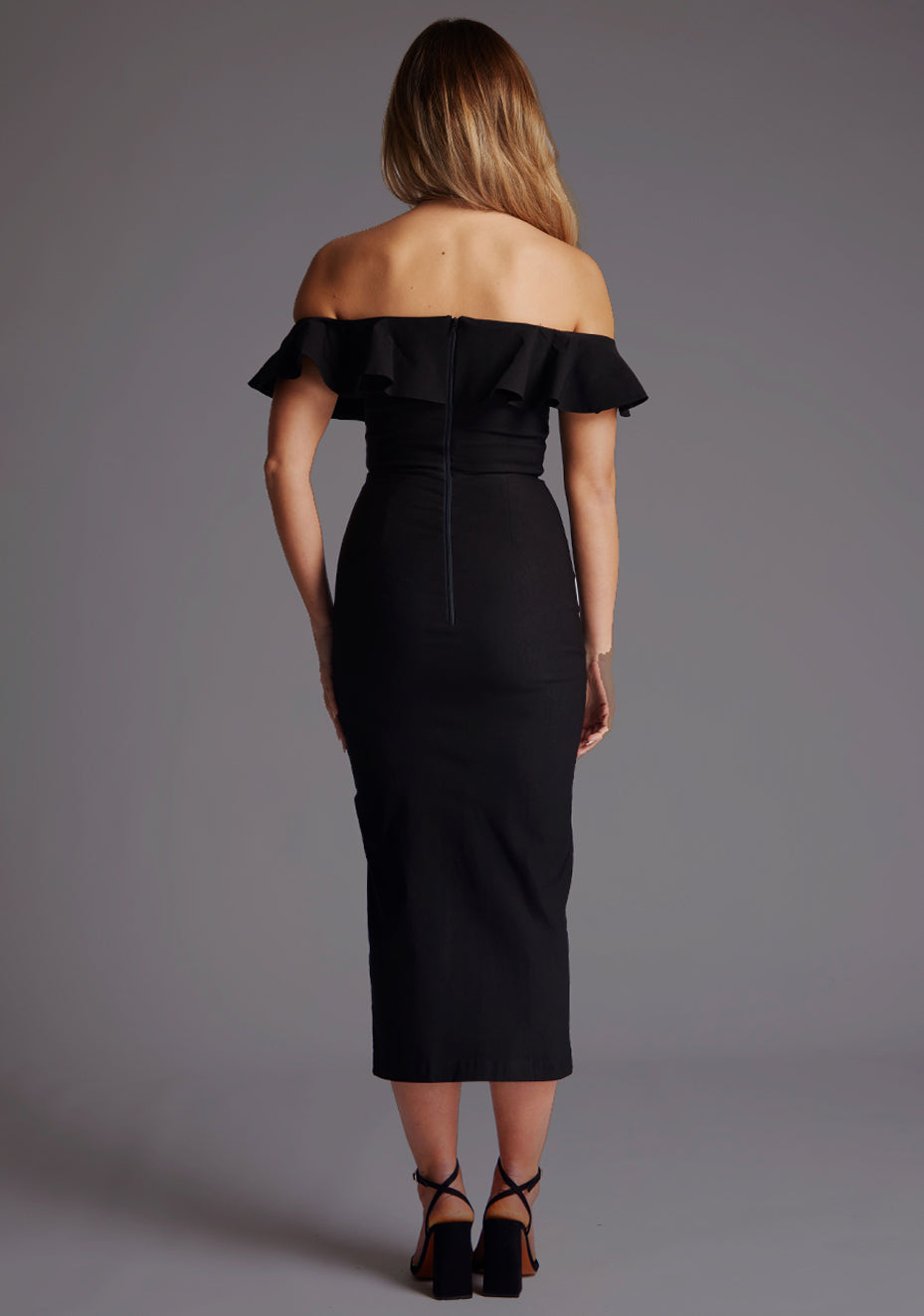 Back image of a model wearing a black midaxi dress with a bardot neckline with a ruffle design, front skirt split and invisible centre back zip. The design featured is the Vesper Deirdre Bardot Off-Shoulder Frill Midaxi Dress in Black.