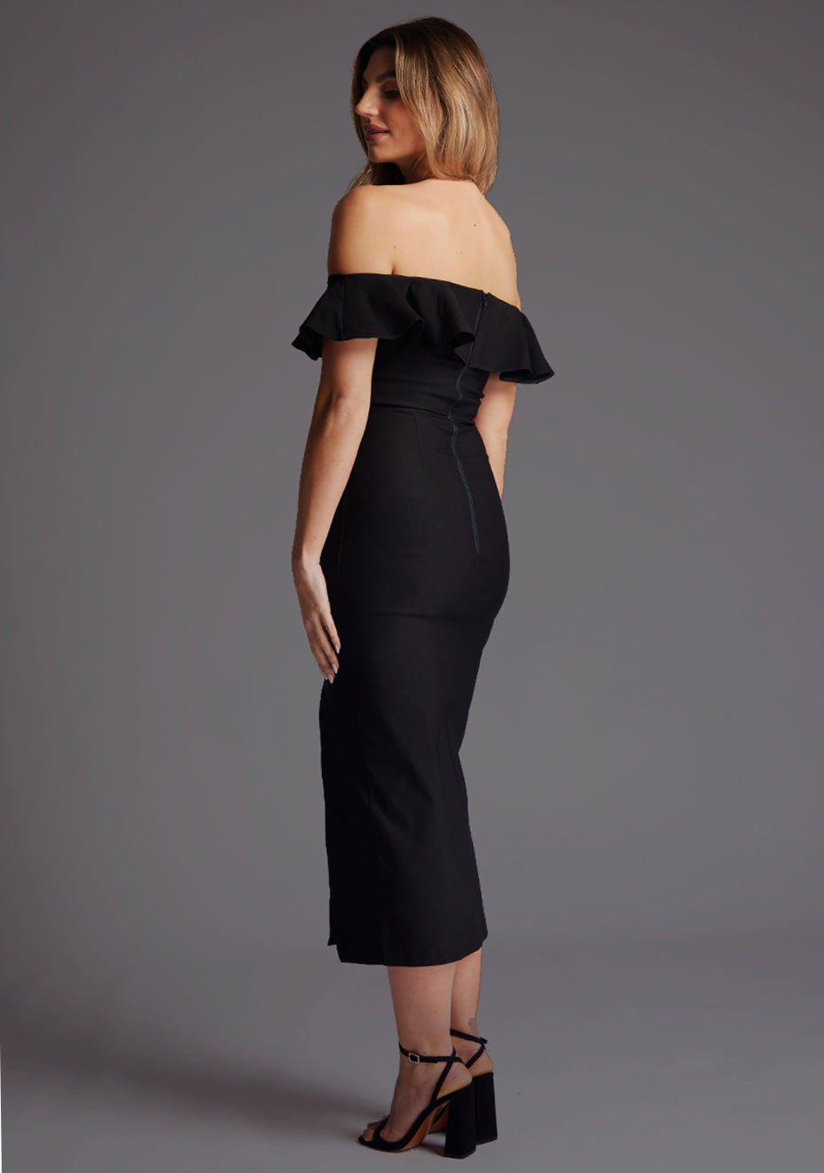 Side back image of a model wearing a black midaxi dress with a bardot neckline with a ruffle design, front skirt split and invisible centre back zip. The design featured is the Vesper Deirdre Bardot Off-Shoulder Frill Midaxi Dress in Black.