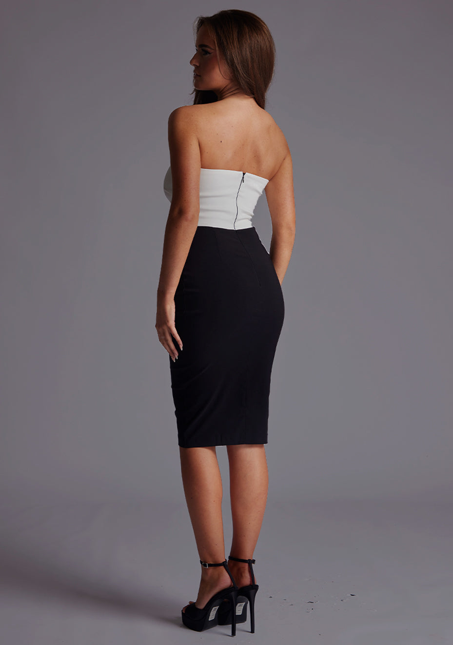 Side backimage of a model wearing a monochrome midi dress with a stunning strapless and the back zipper ensures a comfortable fit. The design featured is the Vesper Drew Strapless Midi Dress in Monochrome.