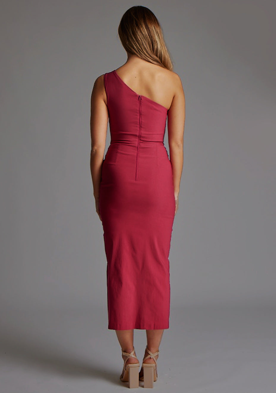 Back image of blonde model wearing a raspberry one shoulder midaxi dress with a asymmetric neckline, one shoulder and front skirt split. The design featured is the  Vesper Gianna One Shoulder Midaxi Dress in Raspberry.