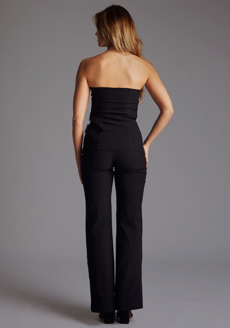 Back image of a model wearing black wide leg trousers with a high-waisted with a thick waistband to accentuate curves. Sleek V-shaped cut-out at the back adds allure. The design featured is the Vesper June Wide Leg Trousers in Black and paired with the VSPR  Sharon Strapless Top in Black.