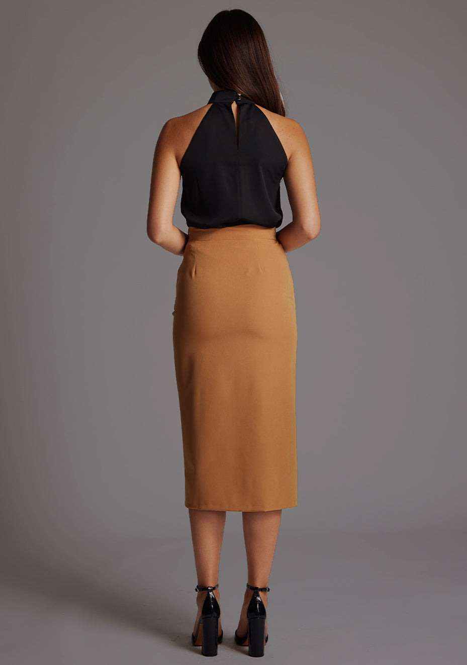 Back image of the model wearing  black top with a  subtle cut-out detail, adding a touch of modern flair to your outfit.  The design featured is the  Vesper Juniper Halter High-Neck Top in Black paired with Sherine Camel Skirt.