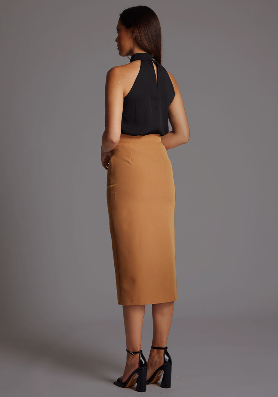 Side back image of the model wearing  black top with a  subtle cut-out detail, adding a touch of modern flair to your outfit.  The design featured is the  Vesper Juniper Halter High-Neck Top in Black paired with Sherine Camel Skirt.