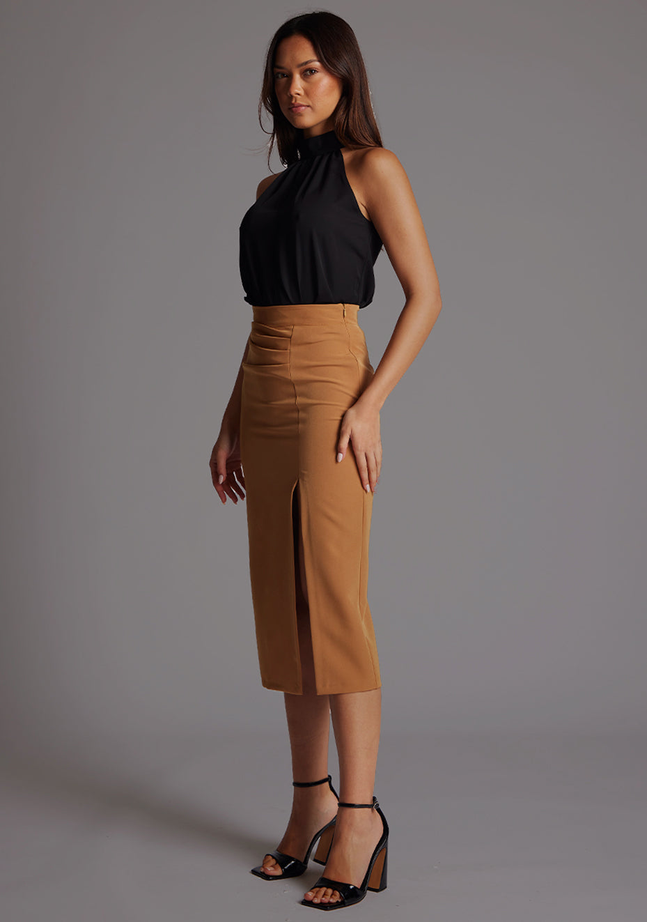 Side front image of the model wearing  black top with a  subtle cut-out detail, adding a touch of modern flair to your outfit.  The design featured is the  Vesper Juniper Halter High-Neck Top in Black paired with Sherine Camel Skirt.