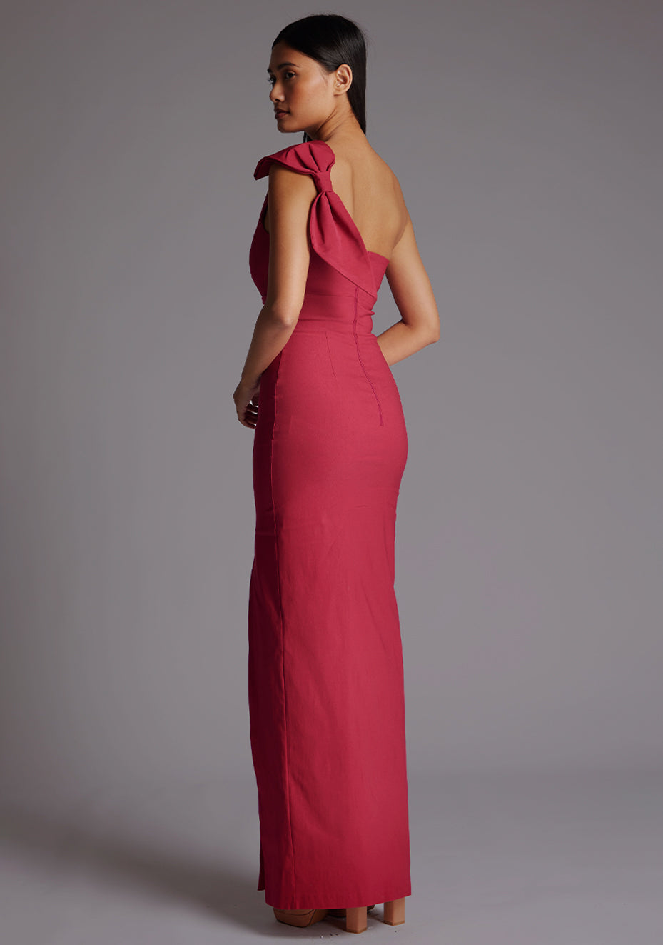 Side back image of a model wearing a raspberry maxi dress, featuring an asymmetric neckline, one shoulder with statement bow detail, and a front skirt split. The dress feature is the  Vesper Keegan One Shoulder Maxi Dress in Raspberry.