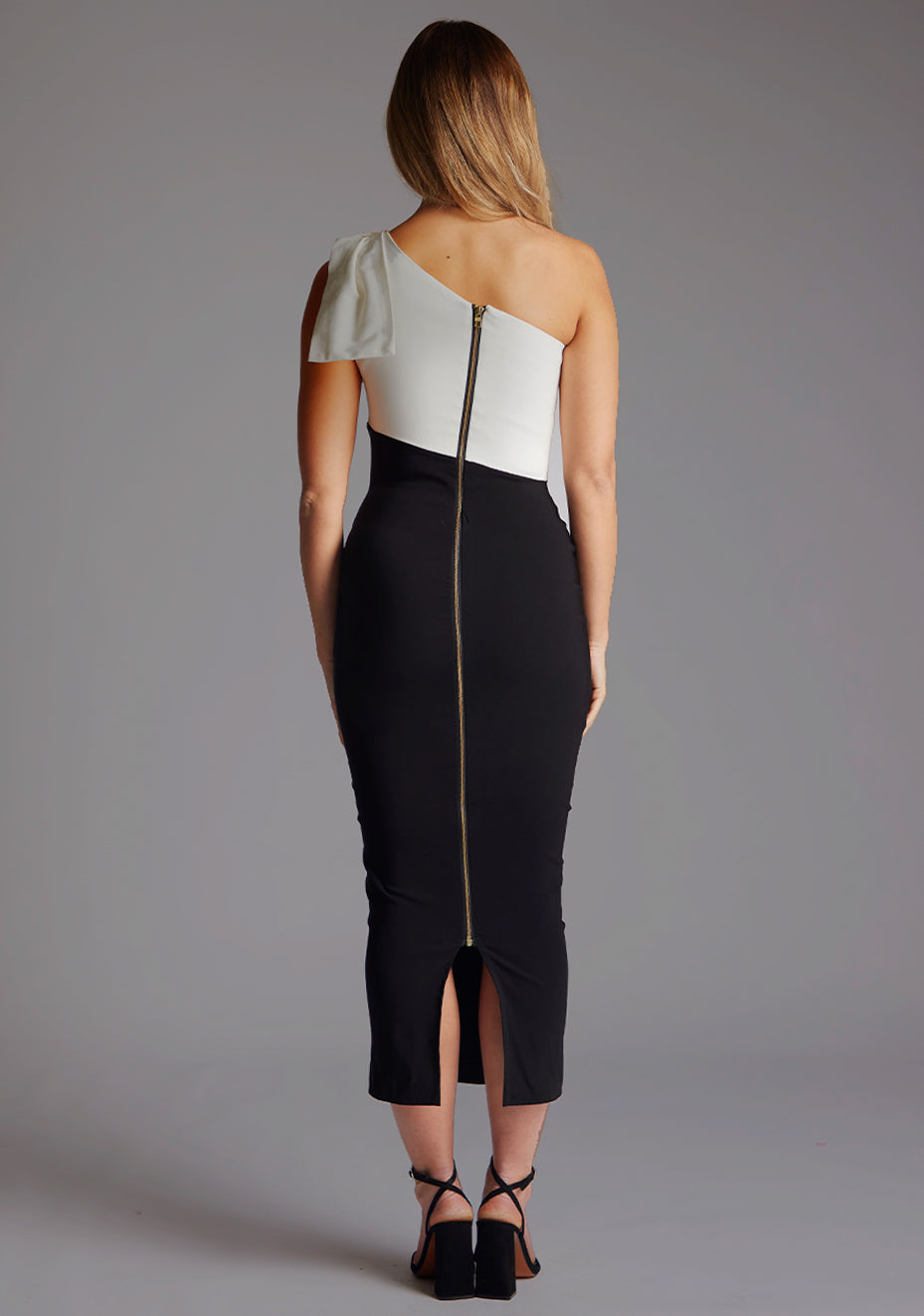 Back image of a model wearing a monochrome one shoulder midaxi dress with a elegant bow on the shoulder, an asymmetric neckline and statement gold zip. The design featured is the Vesper Libby One Shoulder Midaxi Dress in Monochrome.