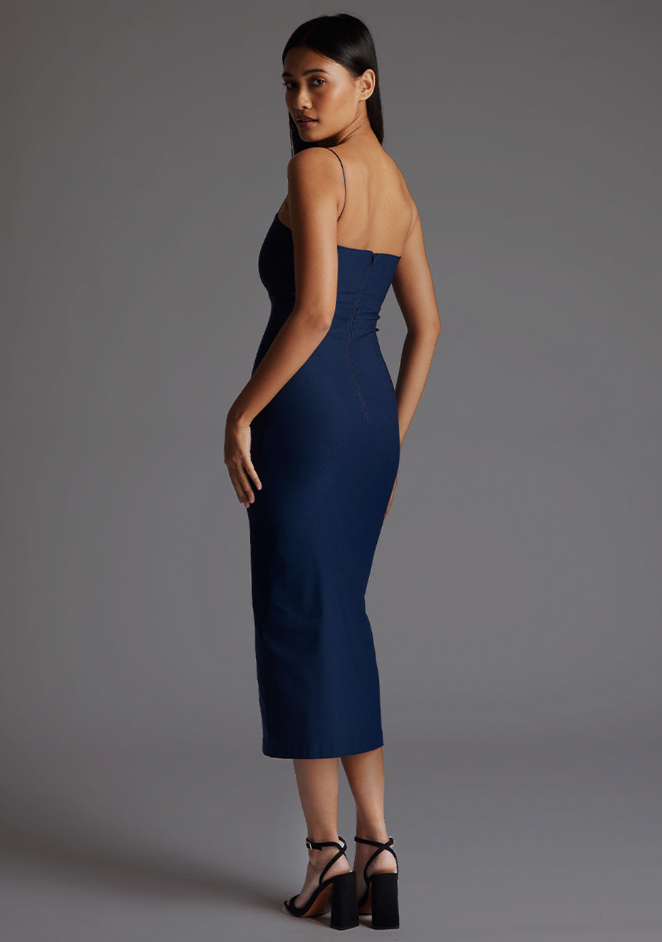 Side back image of  model wearing a navy strappy midaxi dress with a sweetheart neckline, thin straps, centre front skirt split and centre back zip for a perfect fit. The design featured is the Vesper Miren Strappy Bodycon Midaxi Dress in Navy.