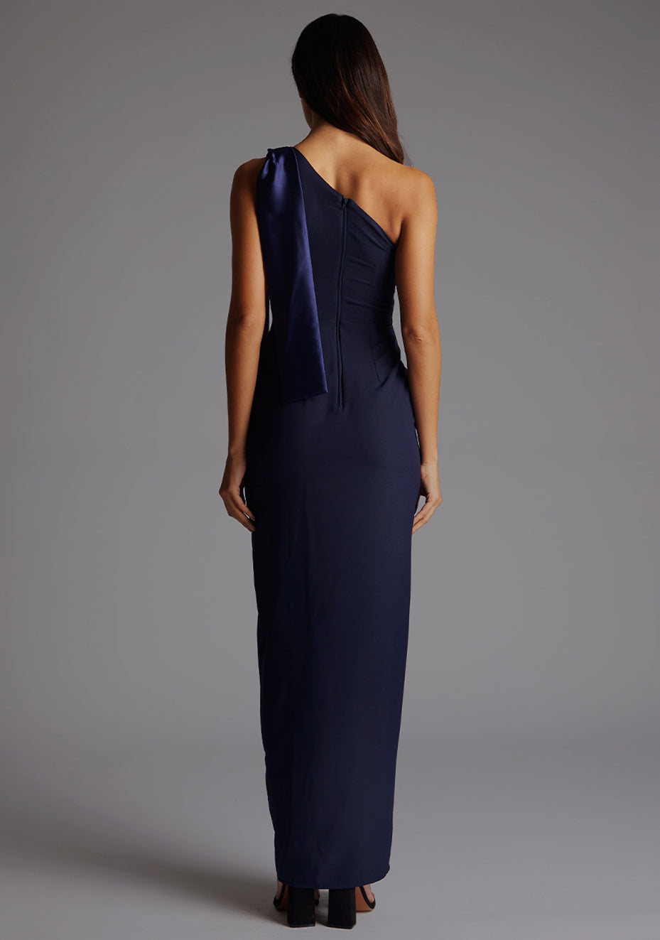 Back image of a model wearing a  One Shoulder Navy Maxi Dress with a  one shoulder contrast satin band and bow detail with a front skirt split. The design features Vesper Olympia One Shoulder Maxi Dress in Navy.