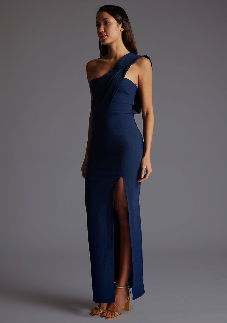 Front quarter image of a model wearing a navy one shoulder maxi dress with a straight neckline with an asymmetric one shoulder draped fabric with pleat detailing and front skirt split. The design featured is the Vesper Marni Navy One Shoulder Maxi Dress