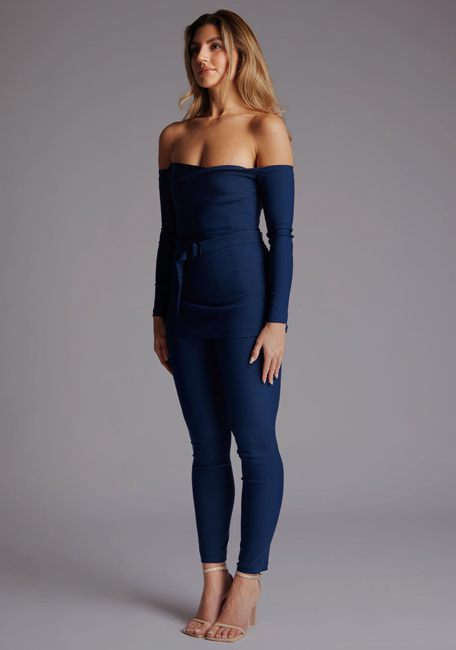 Front quarter  image of a model wearing a navy top paired with matching navy trousers. The top features a bardot neckline with soft sweetheart effect, long sleeves and a belt. The styles featured are Vesper Parisa Navy Long Sleeved Bardot Top and Vesper Danica Navy Trousers 