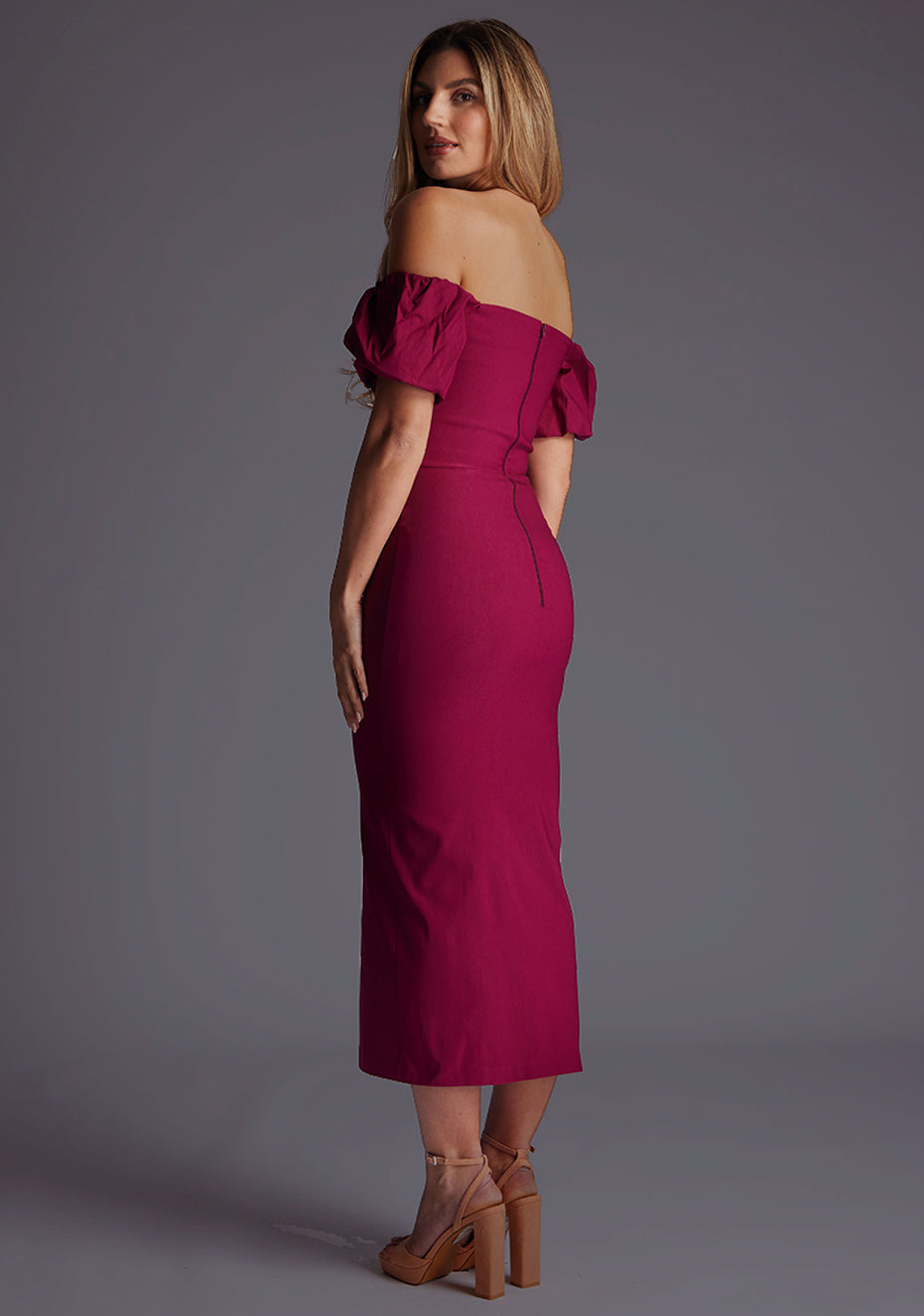 Back quarter image of a blonde model wearing a berry midaxi dress with a bardot neckline gracefully frame off shoulders, while the back zip ensures a seamless silhouette. The design featured is the Vesper Quinton Berry Midaxi Dress.