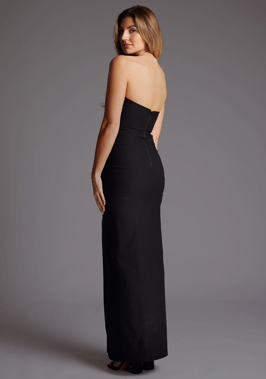 Back quarter image of a blonde model wearing a black bodycon maxi dress with a  strapless elegance with a front skirt split for graceful movement and flattering slim fit enhances silhouette.  The design featured is the Vesper Remington Black Bodycon Maxi Dress.
