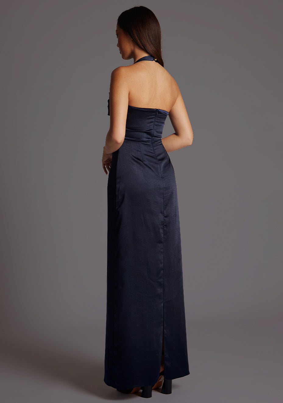 Back quarter image of a model wearing a navy satin maxi dress with a halter cowl neck and low backless, the luxurious satin fabric slim-fit dress effortlessly flatters your figure.  The design featured is the Vesper Saige Navy Satin Maxi Dress.