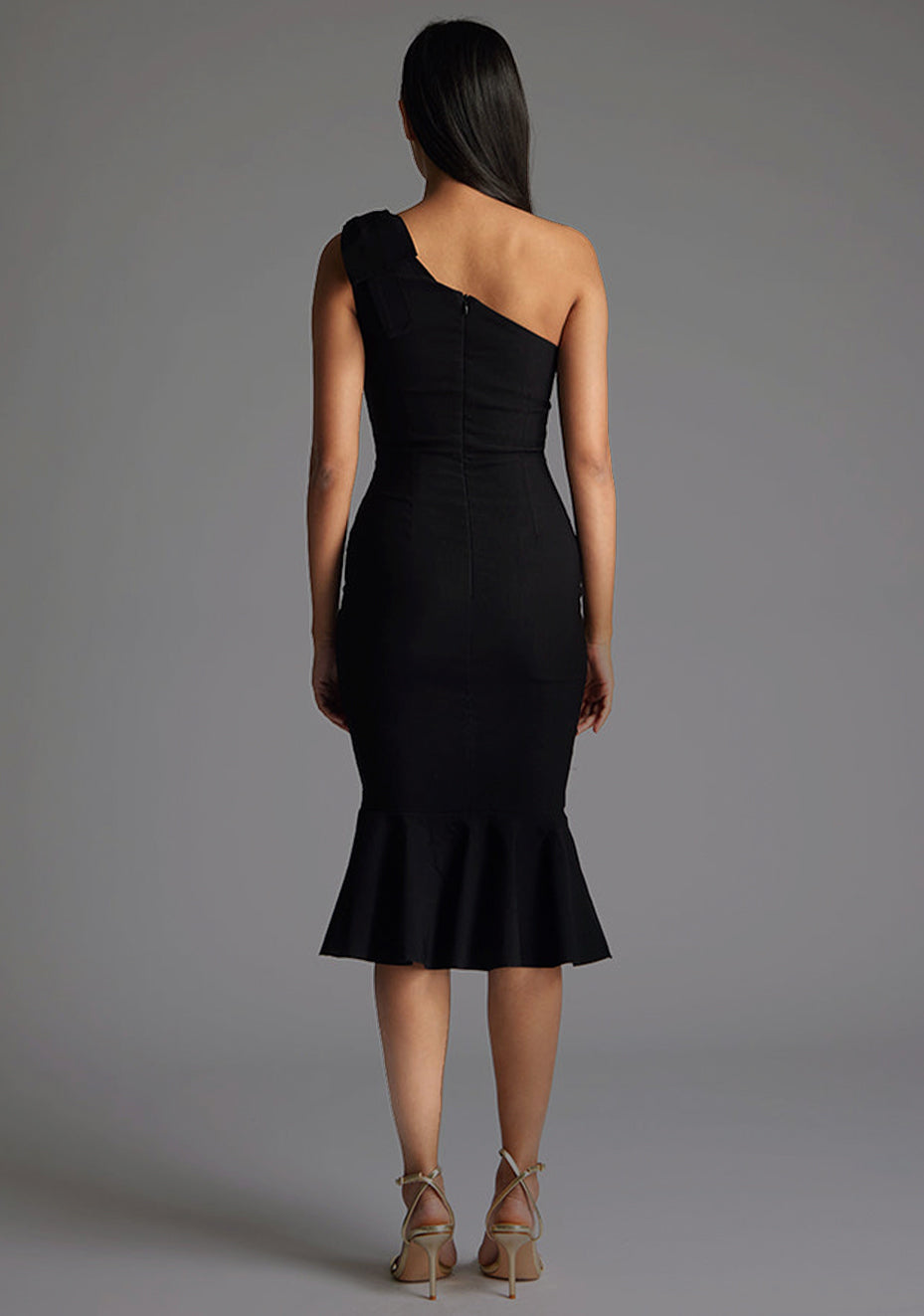 Back image of a model wearing a black one shoulder midaxi dress with a asymmetric neckline, playful one-shoulder with bow detail, and frill hem. The design featured is the Vesper Savannah Black One Shoulder Midaxi Dress.