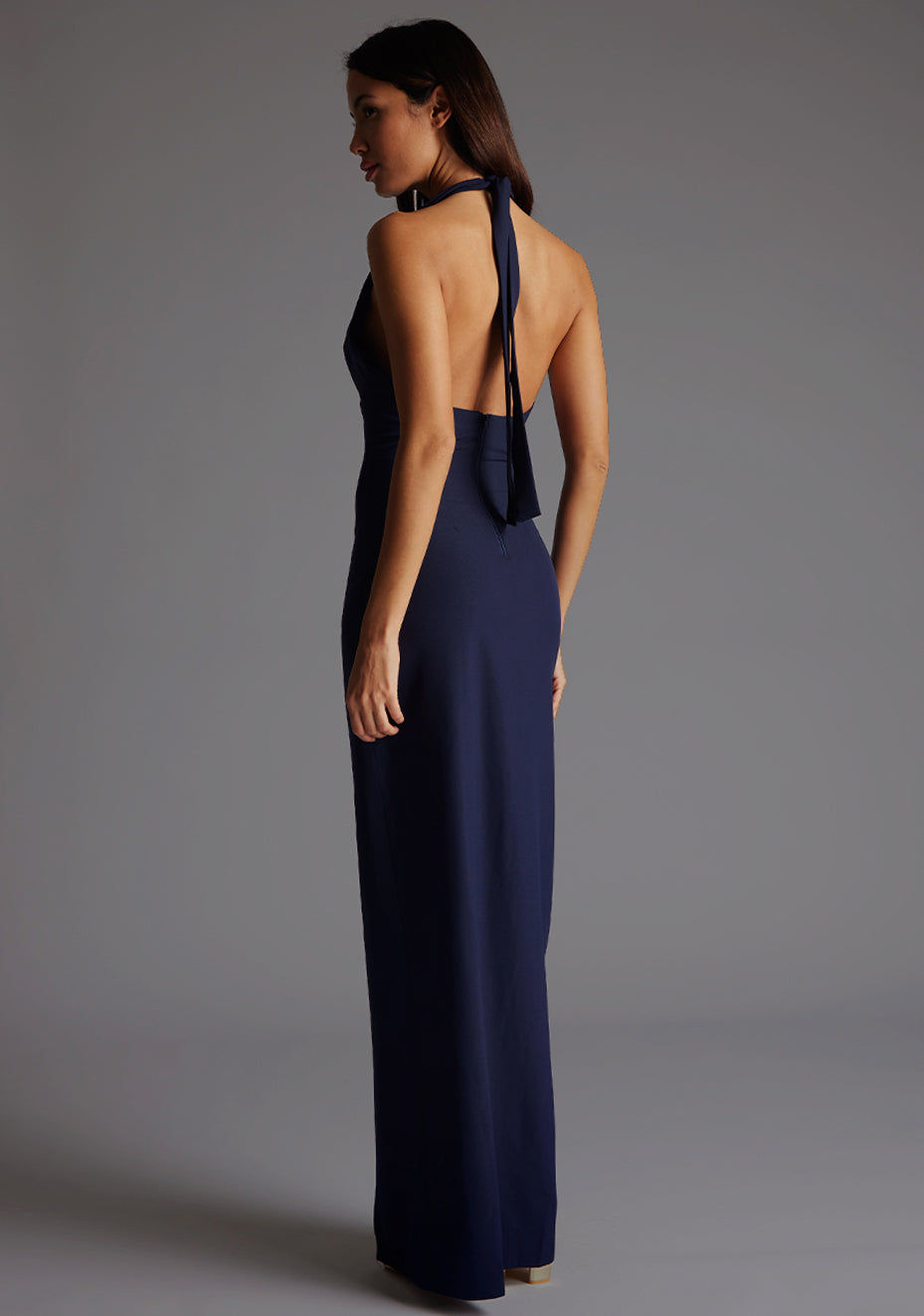 Back quarter  image of a blonde model wearing a navy maxidress with a v-plunging neckline, halter design and a front skirt split. The design featured is the Vesper Sedona Navy Maxi Dress.